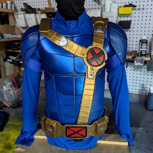 May include: A blue and gold superhero costume, featuring a chest piece with a gold harness and an X-shaped emblem. The costume includes a long-sleeved blue shirt and a gold belt with a red X emblem. The shoulder pads are gold and blue.
