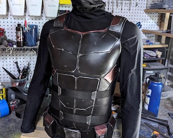Può includere: Un'armatura pettorale nera e grigio metallizzato con accenti rossi, esposta su un manichino. L'armatura presenta un design segmentato con cintura e tasche. Il manichino indossa una maglia nera a maniche lunghe e un cappuccio.
