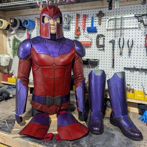 May include: A Magneto costume set featuring a red and purple helmet, a matching chest piece with shoulder armor, forearm guards, two thigh plates, and a pair of tall purple boots. The armor displays a segmented design with a dark red base and purple accents, presented on a mannequin torso with separate leg and foot components.