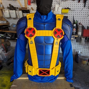 May include: A blue and yellow superhero costume, featuring a chest piece with red and yellow accents and the letter "X". The costume includes a yellow belt with pouches and a matching harness. The costume is displayed on a mannequin.