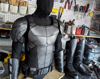 May include: A black tactical costume set featuring a full-face mask, a segmented chest plate with shoulder guards, forearm bracers, a utility belt, and a pair of black boots. The armour pieces have a metallic silver finish with dark weathering.