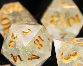 May include: A set of iridescent dice with gold numbers. The dice are made of clear resin with a glittery finish. The dice are scattered on a black fabric background.