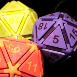 May include: Three twenty-sided dice in bright neon yellow, purple, and coral orange. Each die features triangular faces with clearly printed numbers. The coral die displays numbers 19, 11, and 9, while the purple die shows 18, 15, 5, and 13.