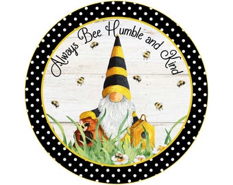 Op de afbeelding: Een rond houten bord met een zwarte en witte stippenrand. Het bord toont een kabouter met een gele en zwarte gestreepte hoed en een bijenkorf in zijn hand. De tekst op het bord luidt "Always Bee Humble and Kind".