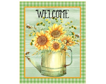 Op de afbeelding: Een groene gieter met een rustieke uitstraling, gevuld met een boeket gele zonnebloemen en groen blad. De achtergrond is een lichtblauw houten paneel met het woord "WELCOME" in zwarte letters. De gieter is omgeven door een groen-wit ruitpatroon.