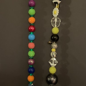 May include: Two colorful beaded strings with a green bow at the top of one and a clear plastic charm at the top of the other. The beads are various colors, shapes, and sizes, including green, pink, blue, yellow, black, and white.