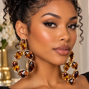 May include: Large drop earrings featuring a series of interconnected, irregular amber-colored resin stones encased in polished gold-tone metal frames. The design consists of five distinct, rounded segments arranged in an open, elongated oval shape.
