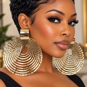 May include: Large gold-toned earrings featuring a series of concentric, hammered-texture circular rings suspended from a smooth, polished rectangular top piece. The metallic design creates a layered, circular effect that hangs prominently.