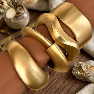 May include: A set of three gold-toned metal bracelets with a brushed matte finish. The collection includes one wide cuff, one wavy-shaped bangle, and one smooth, rounded bangle, all worn together on the arm to create a layered appearance.