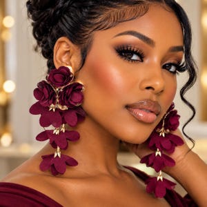 May include: A pair of long statement earrings featuring multiple layers of deep burgundy fabric flower petals attached to gold-toned metal connectors. The floral design cascades downward in a tiered arrangement, creating a bold and textured appearance.
