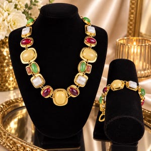 May include: A gold-tone statement necklace and matching bracelet set featuring alternating oval and square cabochon stones in shades of deep red, pale yellow, white, and green, all set within polished gold-colored metal frames and connected by links.