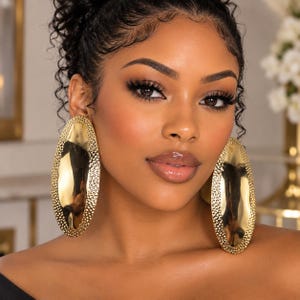 May include: A pair of large, oval-shaped gold earrings featuring a smooth, reflective center and a textured, hammered border. The earrings have a polished metallic finish and a substantial size that frames the face with a bold, shiny appearance.