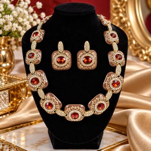 May include: A jewelry set consisting of a necklace and matching earrings. Each piece features square, tan-colored enamel frames with gold-tone filigree borders, centered with deep red, faceted, oval-shaped stones and small clear crystals.