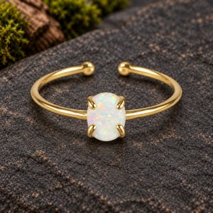 May include: A gold-toned open ring featuring a single oval-shaped iridescent opal gemstone set in a four-prong setting. The band is thin and smooth, with a polished finish that reflects light, showcasing the play of color within the white opal stone.