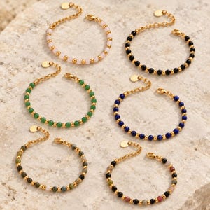 May include: Six gold-tone chain bracelets featuring various gemstone beads. The bracelets include light pink, black, green, blue, and multi-colored stone options. Each bracelet has a gold-tone metal chain, a lobster clasp, and a small round charm.