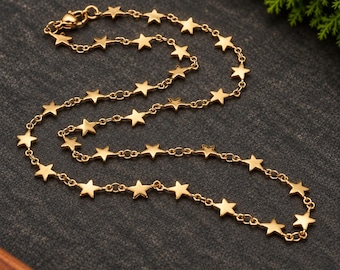 May include: A gold-toned chain necklace featuring a series of evenly spaced, flat, five-pointed star charms linked together. The necklace includes a lobster claw clasp at one end and is arranged in a curved, circular shape on a dark surface.