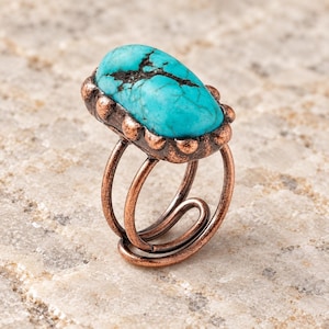 May include: A ring featuring a large, oval-shaped turquoise stone with dark matrix veining, set in a textured, dark copper-toned metal bezel with small bead accents. The band is constructed from a single piece of coiled copper-toned wire.