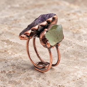 May include: A ring featuring a double-wire copper band with a dark, oxidized finish. The ring is set with two raw, uncut stones: a large, elongated deep purple amethyst and a smaller, angular pale green crystal, both held by copper prongs.