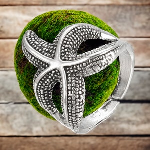May include: A silver starfish ring with a textured surface. The ring is set on a bed of green moss.
