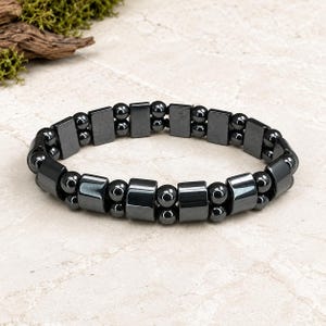 May include: A stretch bracelet featuring a repeating pattern of polished dark gray hematite beads. The design alternates between single rectangular block beads and pairs of small round spherical beads arranged in a continuous circular band.