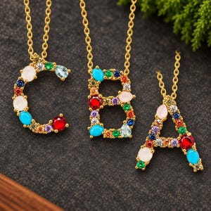 May include: Three gold-tone necklaces featuring letter pendants C, B, and A. Each letter is embellished with an array of multi-colored gemstones including red, blue, green, orange, purple, and white stones in various round and teardrop shapes.