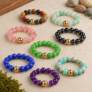May include: A collection of eight elastic bracelets, each featuring a single gold-toned metal bead centered among smooth, round gemstone beads. The bracelets display various colors including pink, green, blue, purple, black, and multi-colored earth tones.