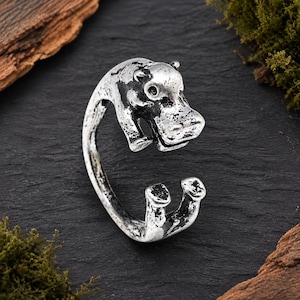 May include: A silver-toned metal ring featuring a sculpted hippopotamus design. The band is open, with the head of the hippopotamus forming one end and the animal's front feet forming the other end. The metal has a distressed, oxidized finish.