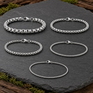 May include: Five silver-toned metal chain bracelets arranged in a group. Each bracelet features a box chain link design with a polished finish and a secure lobster claw clasp. The bracelets vary in thickness, ranging from thicker links to thinner links.