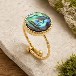 May include: A gold-toned ring featuring a circular setting with an iridescent abalone shell inlay. The band has a textured, hammered finish and an open design, ending in two small gold-toned spheres at the base of the ring's shank.