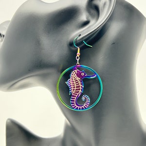 May include: A pair of iridescent rainbow earrings featuring a seahorse design. The earrings are circular with a cutout of a seahorse in the center. The seahorse is a purple color with a gold outline.