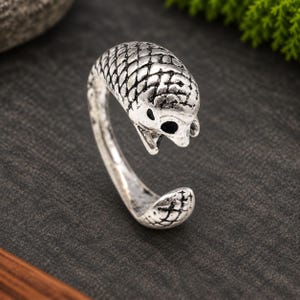 May include: An open-ended silver-toned ring featuring a textured, scale-like pattern across the surface. The design depicts a stylized creature head with dark, recessed detailing in the eyes and mouth area, creating a high-contrast, weathered finish.