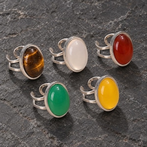 May include: Five adjustable silver-tone rings, each featuring a smooth, oval-shaped cabochon stone. The stones are tiger's eye, opaque white, deep red, bright green, and vibrant yellow. Each ring has a split, open-ended band design.