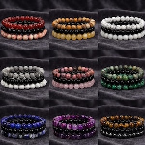 May include: Nine stacks of beaded bracelets in various colors, including red, black, white, and purple. Each bracelet is made of round beads. The bracelets are displayed on a dark gray fabric surface.