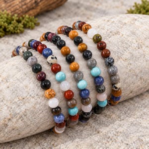 May include: A collection of five elastic bracelets made from round, multi-colored natural stone beads. The beads feature various textures and patterns in shades of blue, red, black, white, orange, grey, and brown, arranged in repeating sequences.