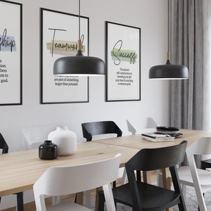 May include: Three black framed prints with white text on a white wall. The prints feature the words "Leadership", "Teamwork", and "Success" with definitions below each word. The prints are hanging above a wooden table with six chairs. Two black pendant lights hang above the table.