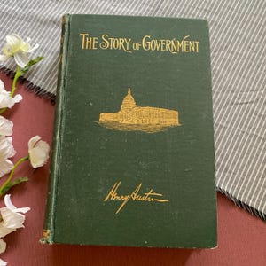 Può includere: Un libro vintage con copertina rigida intitolato "The Story of Government" con scritte dorate e un'illustrazione di un edificio. Il libro ha una copertina verde scuro e il nome dell'autore, Henry Austin, è anch'esso in oro.