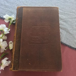 May include: A vintage, brown leather-bound book with a faded embossed design on the cover. The book has a worn appearance, with visible wear on the edges and corners. The background includes white flowers and a striped fabric.