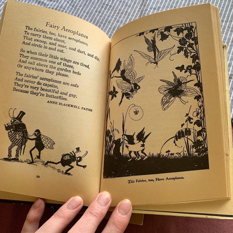 Pu&ograve; includere: Libro aperto con illustrazioni e testo. La pagina sinistra presenta una poesia intitolata "Fairy Aeroplanes" di Anne Blackwell Payne. La pagina destra presenta illustrazioni in bianco e nero di fate e farfalle. Il libro &egrave; aperto su una superficie giallo chiaro.