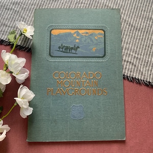 Puede incluir: Una cubierta de libro de color verde salvia con el título Colorado Mountain Playgrounds en letras doradas. La portada presenta una ilustración rectangular de personas a caballo en un paisaje montañoso y un pequeño emblema Union Pacific System.