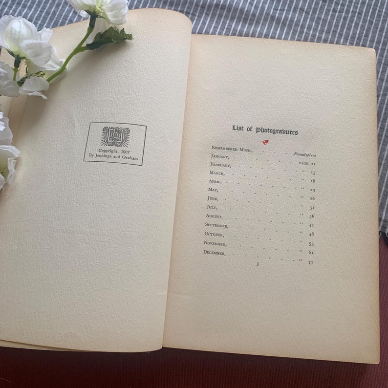Puede incluir: Un libro abierto con p&aacute;ginas envejecidas de color crema. La p&aacute;gina izquierda tiene un emblema impreso y el texto: "Copyright, 1907 By Jennings and Graham". La p&aacute;gina derecha enumera "List of photograbures" con meses y n&uacute;meros de p&aacute;gina. Flores blancas descansan sobre el libro.