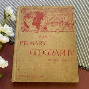 Può includere: Un libro vintage intitolato "Frye's Primary Geography, General Edition" di Ginn & Company. La copertina presenta un'illustrazione rossa di una mappa del mondo e navi che navigano sul mare.