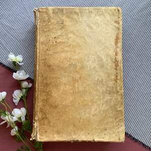 May include: A vintage, worn, tan-coloured book with a textured cover. The book is lying flat on a striped fabric surface.