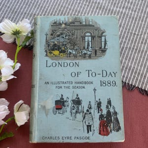 Puede incluir: Un libro antiguo titulado "London of To-Day" de 1889. La portada es de color azul claro con ilustraciones de carruajes tirados por caballos y personas. El título y el autor, Charles Eyre Pascoe, son visibles.
