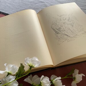 Peut inclure: Un livre ouvert avec un dessin au crayon d'un groupe d'anges sur la page de droite. La page de gauche est vide. Des fleurs blanches sont au premier plan.
