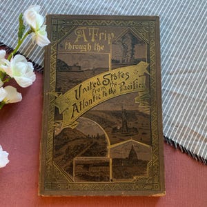 Puede incluir: Un libro antiguo titulado "A Trip Through the United States from the Atlantic to the Pacific." La portada presenta un fondo marrón con letras doradas e ilustraciones de varios monumentos.