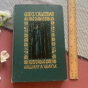 Puede incluir: Un libro antiguo de tapa dura titulado "God's Calendar" de William A. Quayle. La portada es de color verde oscuro con letras doradas y una ilustraci&oacute;n de un bosque. Una regla de madera se coloca junto al libro, midiendo unos 25 cm.