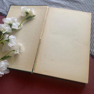 Puede incluir: Un libro abierto con p&aacute;ginas envejecidas de color crema. Flores blancas con tallos verdes est&aacute;n dispuestas en el lado izquierdo del libro. La cubierta del libro es de color verde oscuro y la encuadernaci&oacute;n es visible.