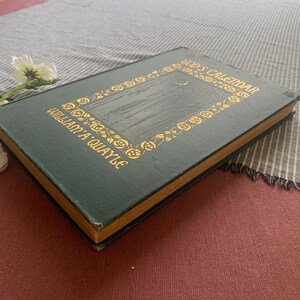 Puede incluir: Un libro antiguo titulado "God's Calendar" de William A. Quayle. El libro tiene una cubierta verde oscuro con letras doradas y un borde floral decorativo. El libro est&aacute; sobre una tela a rayas.