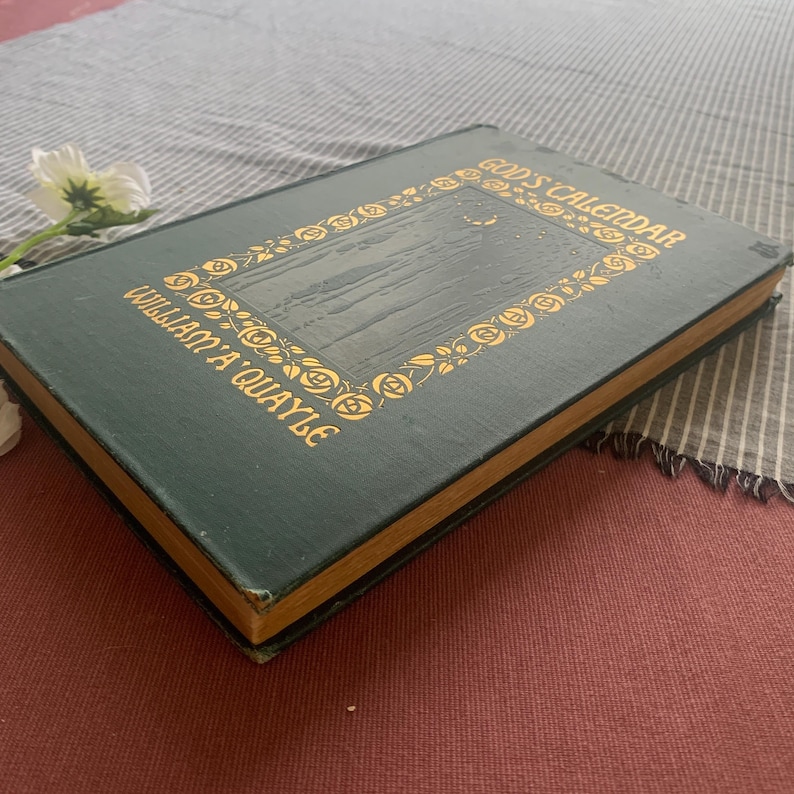 Puede incluir: Un libro antiguo titulado "God's Calendar" de William A. Quayle. El libro tiene una cubierta verde oscuro con letras doradas y un borde floral decorativo. El libro est&aacute; sobre una tela a rayas.