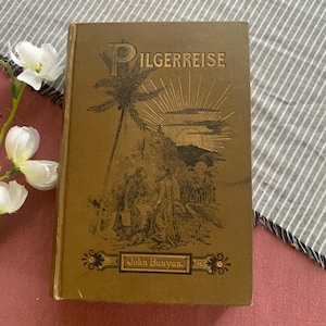 May include: An antique book titled "Pilgerreise" by John Bunyan. The cover is a muted gold color with an illustration of people and a palm tree. The title is in a stylized font, and the author's name is at the bottom.